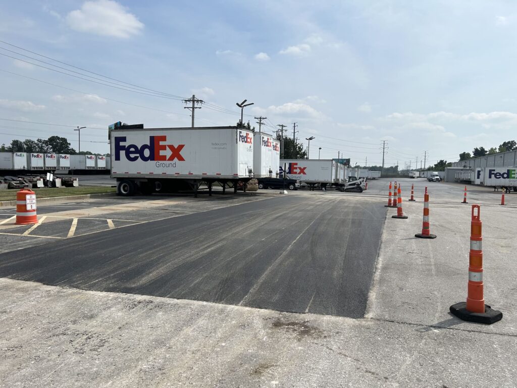 Asphalt parking lot paving project at a FedEx facility, featuring freshly paved surface, parked FedEx trucks, and traffic cones marking the area.