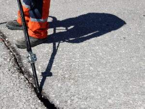Worker applying hot crack sealant to a large asphalt crack using a wand applicator, wearing orange safety pants and black boots on a textured pavement surface.