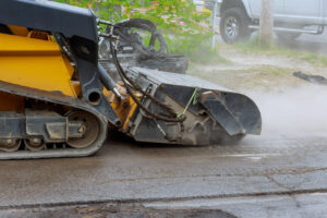 Close-up of an asphalt milling machine removing the top layer of pavement during road maintenance work, with visible dust and debris around the equipment.