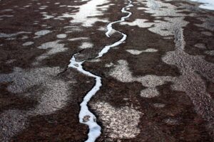 Cracked asphalt pavement with melting snow and ice inside the split, showing frost heave damage caused by winter freeze–thaw cycles.